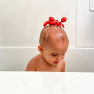 Toddler in bathtub with red rubber crab toy on his head.
