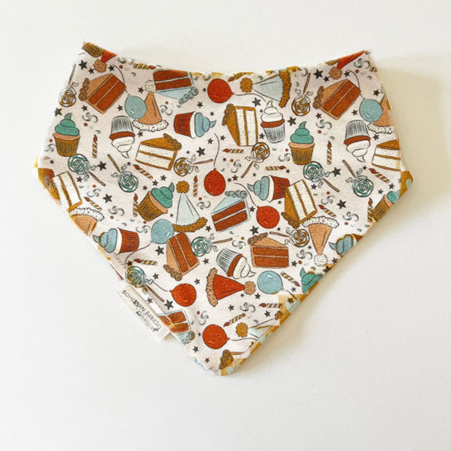 Bohemian Babies made in the USA organic cotton bandana bib. Snap closure. Birthday surprise blue print. Cupcakes, cakes, balloons and lollipops in shades of aqua, brown, mustard, blue and red on an off-white background. Front view.