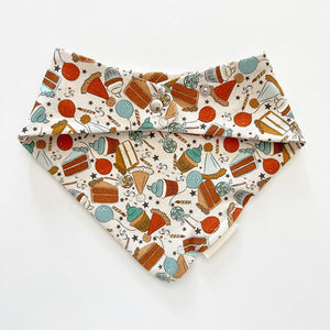 Bohemian Babies made in the USA organic cotton bandana bib. Snap closure. Birthday surprise blue print. Cupcakes, cakes, balloons and lollipops in shades of aqua, brown, mustard, blue and red on an off-white background. Back view.