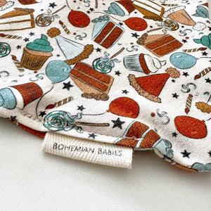 Bohemian Babies made in the USA organic cotton bandana bib. Snap closure. Birthday surprise blue print. Cupcakes, cakes, balloons and lollipops in shades of aqua, brown, mustard, blue and red on an off-white background. Close-up view.