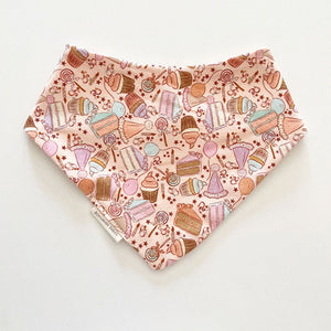 Bohemian Babies made in the USA organic cotton bandana bib. Snap closure. Birthday surprise pink print. Cupcakes, cakes, balloons and lollipops in shades of aqua, brown, peach and lavender on a pink background. Front view.