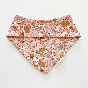 Bohemian Babies made in the USA organic cotton bandana bib. Snap closure. Birthday surprise pink print. Cupcakes, cakes, balloons and lollipops in shades of aqua, brown, peach and lavender on a pink background. Back view.