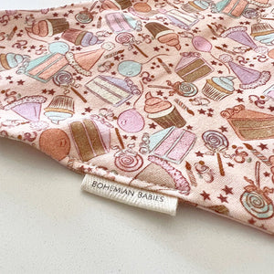 Bohemian Babies made in the USA organic cotton bandana bib. Snap closure. Birthday surprise pink print. Cupcakes, cakes, balloons and lollipops in shades of aqua, brown, peach and lavender on a pink background. Close-up view.