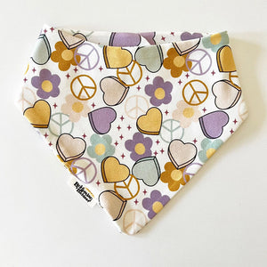 Bohemian Babies made in the USA organic cotton bandana bib. Snap closure. Peace & Love print. Aqua, mustard and purple hearts, peace signs and flowers on an off-white background. Front view.