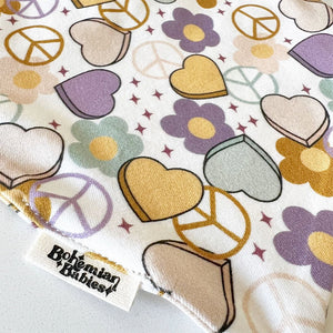 Bohemian Babies made in the USA organic cotton bandana bib. Snap closure. Peace & Love print. Aqua, mustard and purple hearts, peace signs and flowers on an off-white background. Close-up view.