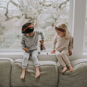 Lifestyle pic of two small children sitting in window at back of a couch. They are both wearing Fox and Poppy pullovers and matching lounge pants. Child on left is in light heather grey; child on right is wearing oat.