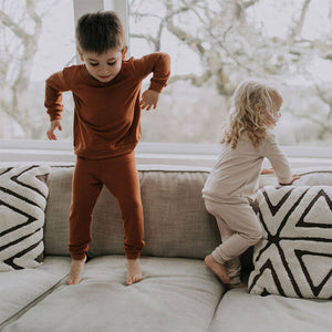 Lifestyle pic of two small children jumping on a couch in front of a window. They are both wearing Fox and Poppy pullovers and matching lounge pants. Child on left is in rust outfit; child on right is wearing oat.