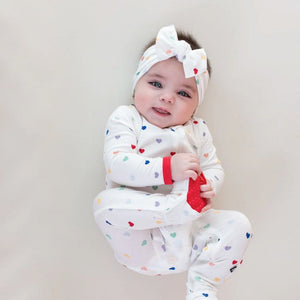 Lifestyle pic of baby with Cloud Rainbow Heart printed bow in her hair wearing Cloud Rainbow Heart printed zip footie with Cardinal Red trim at cuffs and on bottom of non-slip feet.