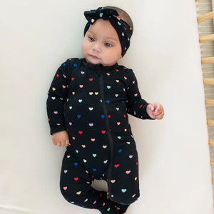Lifestyle pic of baby with Midnight Rainbow Heart printed bow in her hair wearing Midnight Rainbow Heart printed zip footie with Midnight black trim at cuffs and on bottom of non-slip feet.