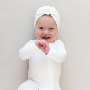 Lifestyle pic of a baby wearing a Cloud white ribbed zippered footie and a Cloud white ribbed headwrap.