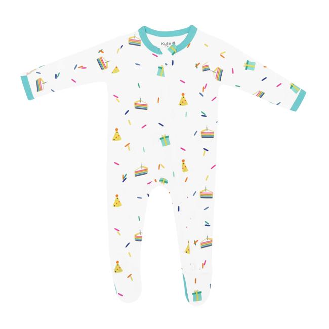 Kyte Baby infant zippered footie with multicoloured cloud party print on a Cloud white background. Zipper down front and one leg with protective zip guard at neck. Robin aqua blue trim at cuffs and neck and on bottoms of non-slip feet over size 6-12m. Cloud Party is a colorful, all-over print featuring rainbow layered cake slices with a lit candle, gift boxes, polka dot party hats, and scattered sprinkles in a rainbow of colors over a white background.