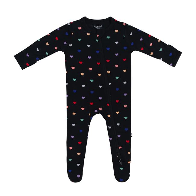 Kyte Baby infant zippered footie with multicoloured tiny hearts on a Midnight black background. Zipper down front and one leg with protective zip guard at neck. Midnight black trim at cuffs and neck and on bottoms of non-slip feet. Midnight Rainbow Heart print is a colorful, all-over print featuring small hearts in Cardinal, Terracotta, Honey, Wasabi, Ice, Royal, and Taro over a black background.